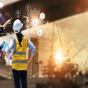 Two construction workers stand to the left side of the image facing a construction site, analyzing data on a tablet while pointing toward project