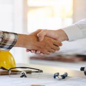 Two individuals shake hands. Behind them is a table with a yellow hard hat on it. 