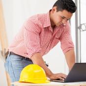 A young construction worker types on a laptop. A yellow hard hat sits off to the side.