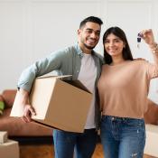 A couple stands in their new house surrounded by moving boxes. One person carries a cardboard box, while the other person holds up the house key.