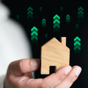 Hand holding wooden house with upward green arrows to indicate increased green building practices