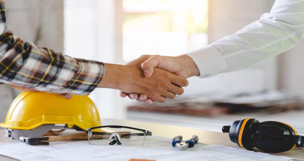 Two individuals shake hands. Behind them is a table with a yellow hard hat on it. 