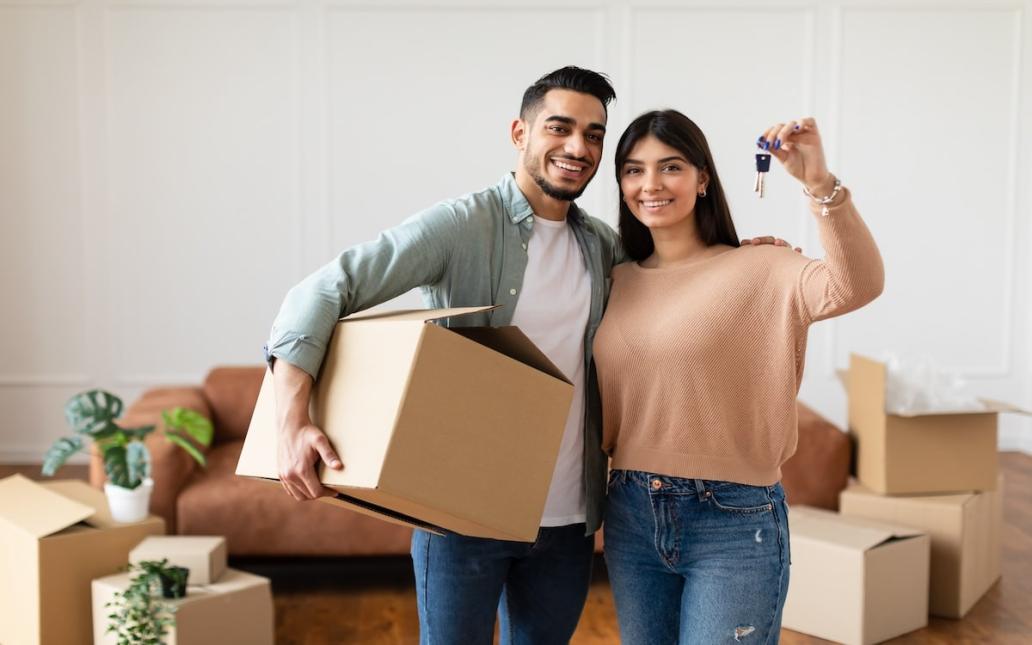 A couple stands in their new house surrounded by moving boxes. One person carries a cardboard box, while the other person holds up the house key.