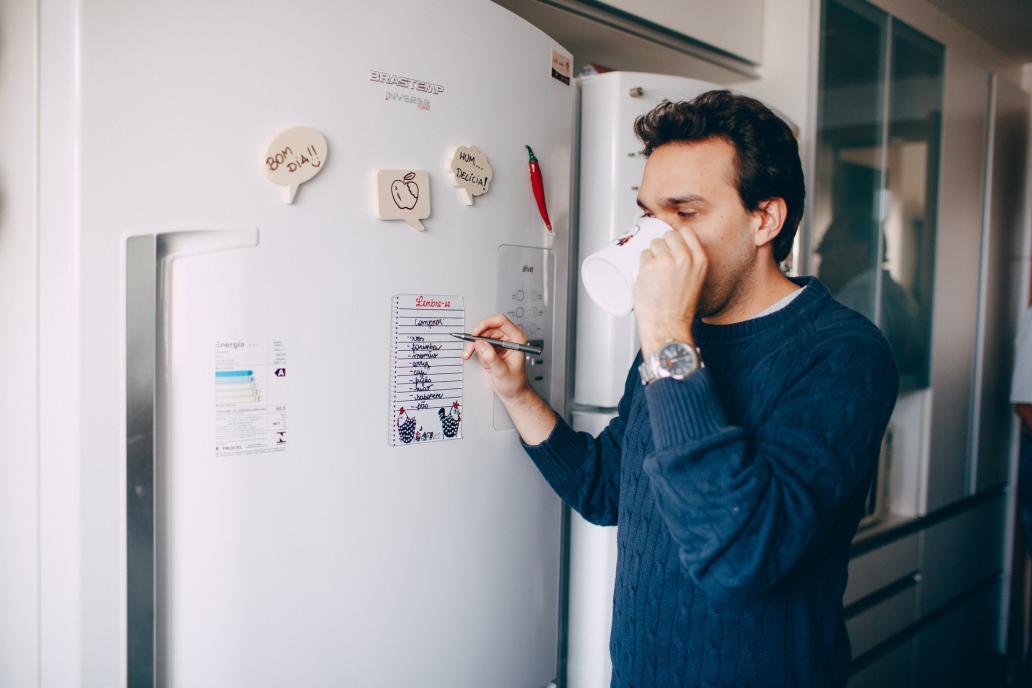 Young man writing reminder on fridge and drinking coffee at home