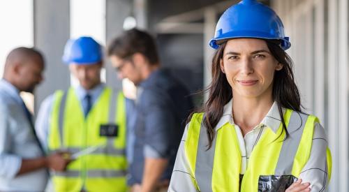 Woman in blue hard hat and yellow construction stands in foreground, while three men stand together in background looking over documents