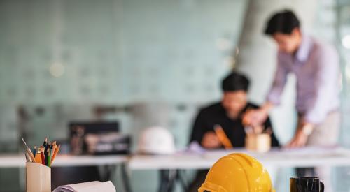 A hard hat and rolled up documents sit on a table in foreground with two individuals in the background working on plans