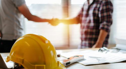 Two construction workers shake hands behind a table that has documents and a yellow hard hat on it