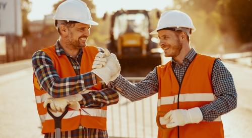 Two construction workers in orange vests and white hard hands shake hands while smiling