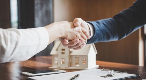 Two people shake hands over paperwork, wooden model home sits on the table in the background