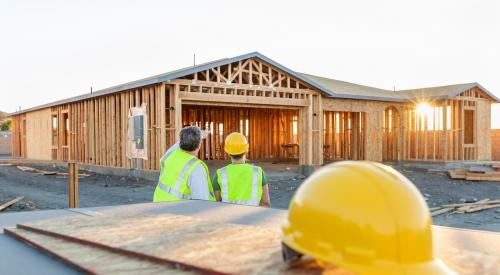 Two construction workers stare off at newly framed house, hard hat sits on plywood behind them