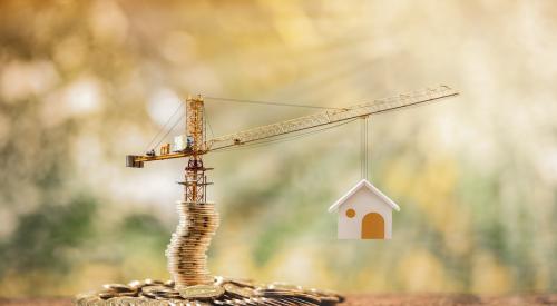 A crane hoisting up a model home sits on a stack of gold coins, indicating the construction loans needed to build housing.