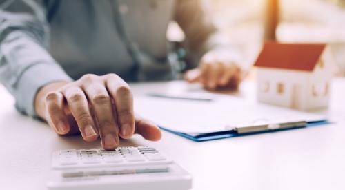 A man sits at a desk, typing on a calculator and looking over documents. A model home sits in the background.