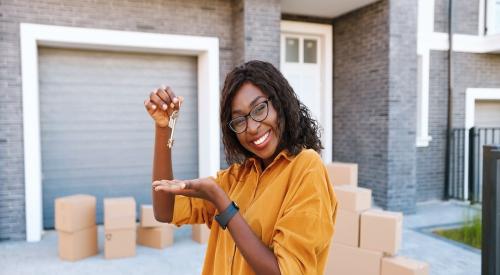 A woman stands in front of a house while smiling and holding up house keys. There are boxes in the driveway, indicating that she is moving in.