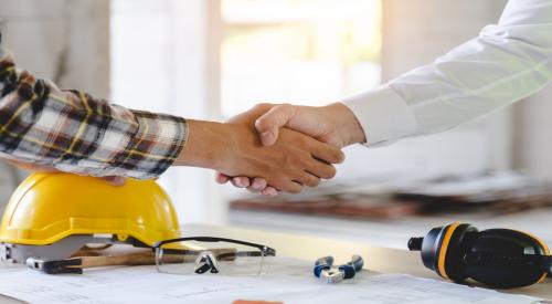 Two individuals shake hands. Behind them is a table with a yellow hard hat on it. 