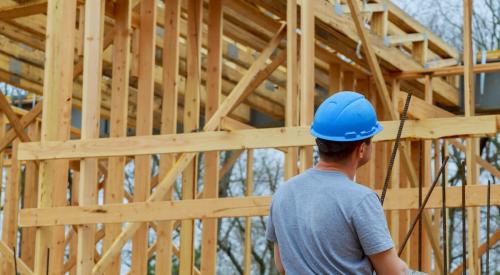 A construction worker in a blue hard hat looks over at home that is under construction