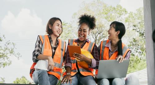 Three women in orange construction vest sit together while smiling and laughing.