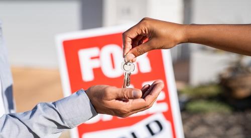 One person hands another person a house key. Behind them is a sign reading "sold," indicating the sale of a home. 