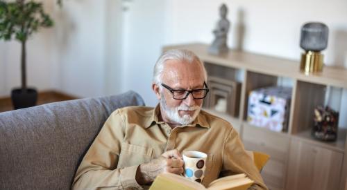 An older man sits on a couch with a cup of coffee and a book