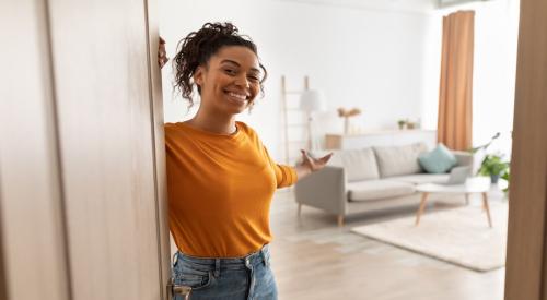 A young woman stands at the door of her home, holding it open to show off the front room