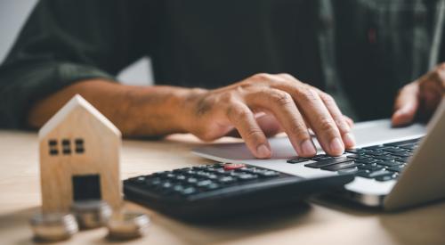 A homeowner works on a laptop. Next to them on a desk are a wooden model house and a calculator.