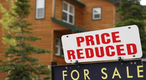 A "For Sale" sign with a "Price Reduced" sign added above it sit in front of an older home