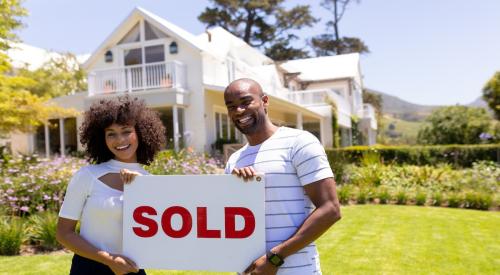 A couple stands in front of their newly purchased home, smiling and holding up a sign that reads "Sold."