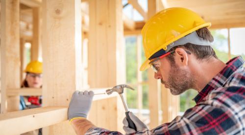 A man in a yellow hard hat hammers a nail into the wall of a home