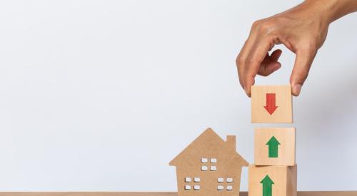 A hand places a wooden block with a down arrow on top of two blocks that have upward arrows. The stack of cubes stands next to a wooden model house.