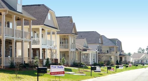 Pictured is a row of homes, and one home has a "for sale" sign in the front yard with a secondary sign reading "reduced."