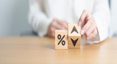 Two wooden blocks sit on a table. One block has a percentage symbol, and the other has an up and down arrow. A hand moves the second block to show the "up" arrow, symbolizing an increase in rates.