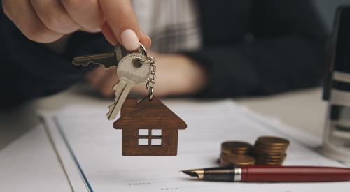 A real estate agent hands over a house key to a buyer. On the desk where they are seated, there is a contract with a pen, indicating it was recently signed.