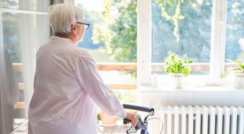 An elderly woman stares out the window of her home