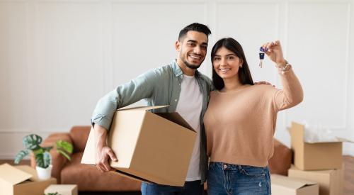 A couple stands in their new house surrounded by moving boxes. One person carries a cardboard box, while the other person holds up the house key.