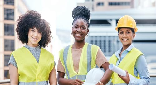 Three young women stand at a construction site wearing yellow vests 