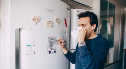 Young man writing reminder on fridge and drinking coffee at home