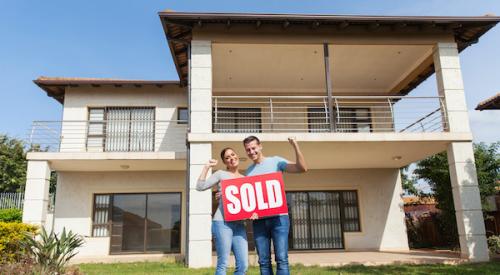 Smiling couple holding 'sold' sign while pointing to new home