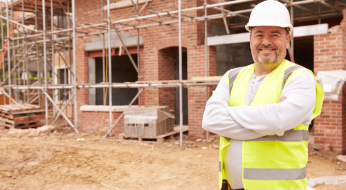 Happy home builder who enjoys his work standing in front of brick home under construction