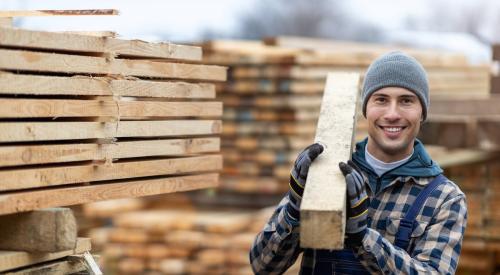 A man is holding a board of lumber with stacks of lumber behind him.