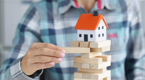 A man is taking a block out of a jenga tower with a mini house on top