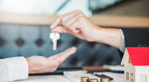 Realtor handing keys to new buyer next to house model and stacks of coins on desk