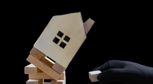 Person with black glove pulling Jenga block out of tipping tower topped by model house