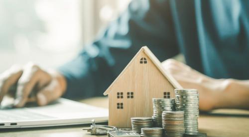 Person calculating costs in front of small wooden model home and stacks of coins