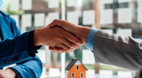 Home buyer and seller shake hands with orange model house and paperwork on the desk in the background