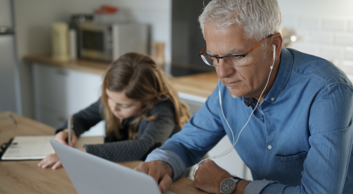 man wearing earbuds working on laptop in kitchen with girl nearby writing at same table