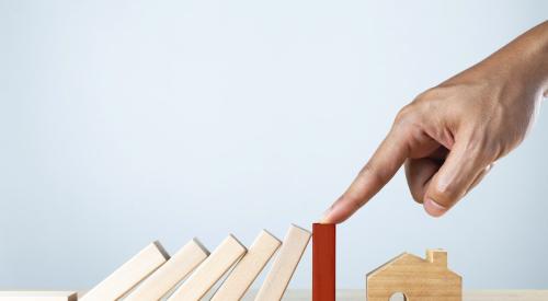 Hand stopping toppling dominoes headed toward small wooden house