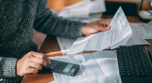 Person sitting at kitchen table using calculator surrounded by utility bills