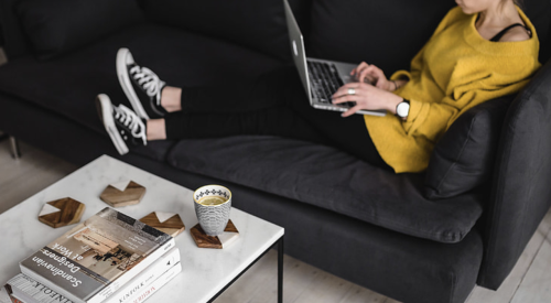 A young woman sitting at home on sofa works on her laptop with a coffee on the table